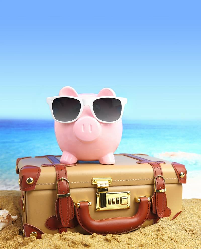 Suitcase with piggy bank in sunglasses on tropical beach